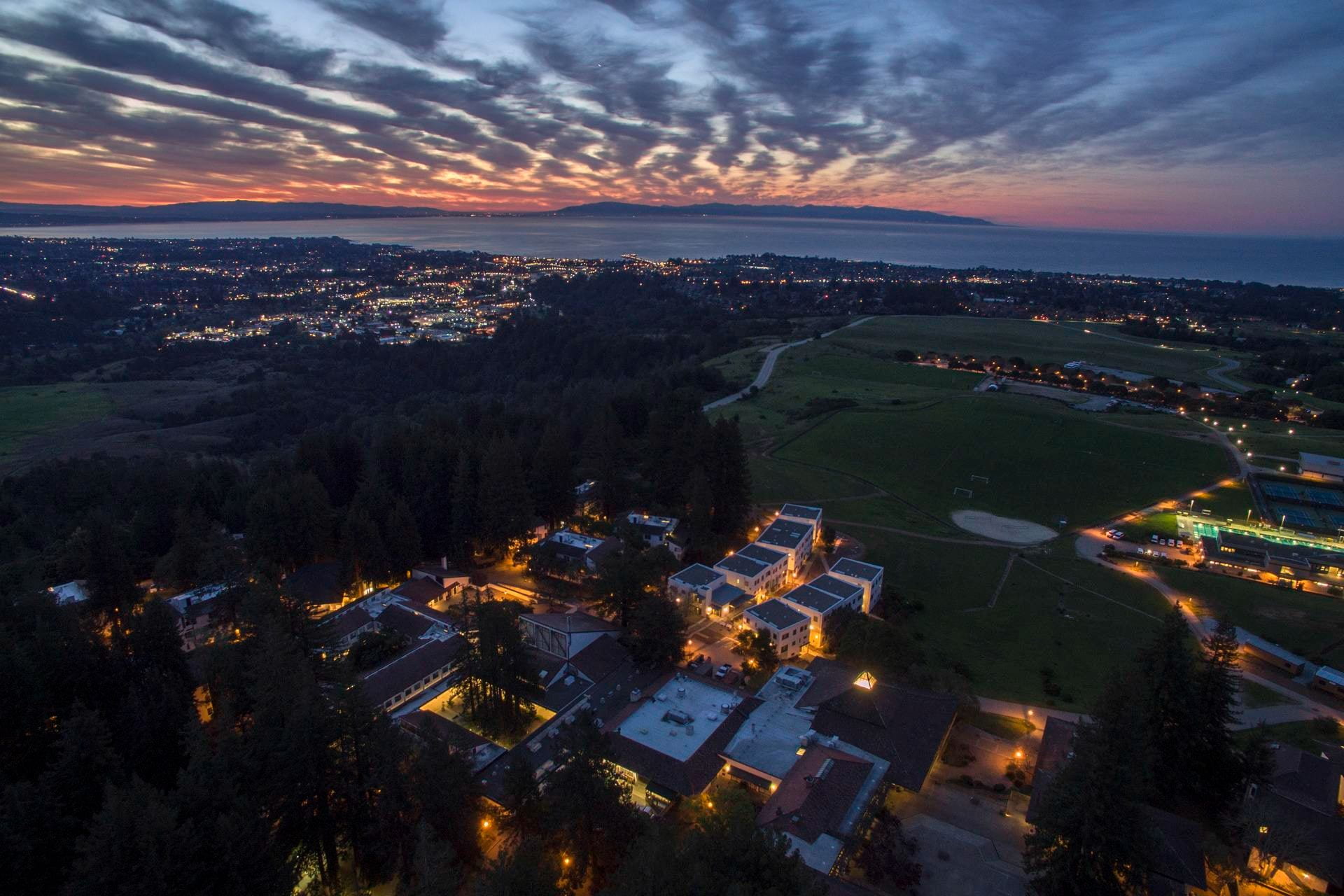 An aerial photo of the UCSC campus at sunset with cloud cover and campus buildings lit up.